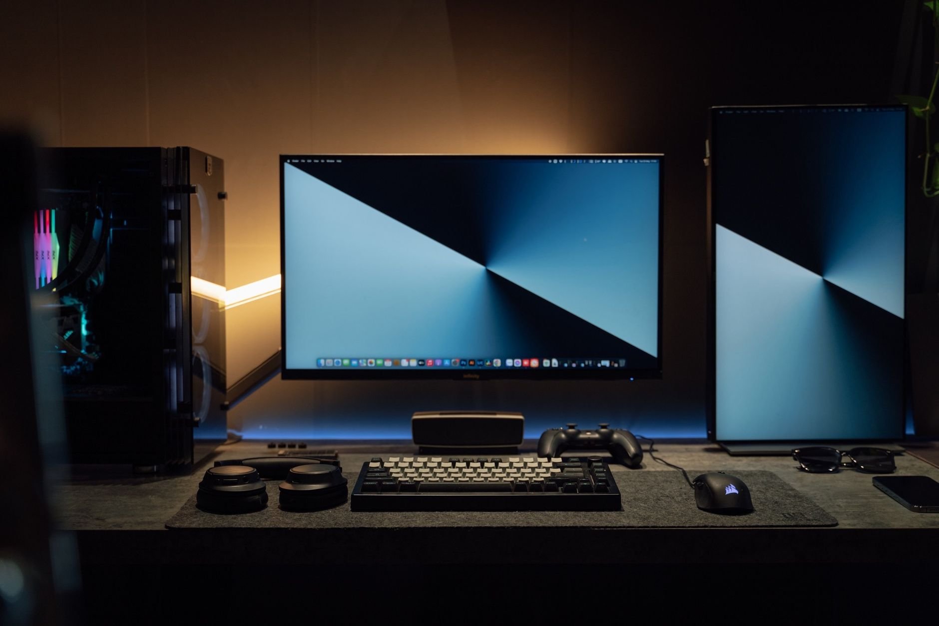 A dimly lit modern computer desk setup featuring dual monitors, a keyboard, and accessories
